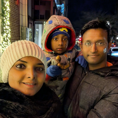 A family selfie taken at night on a city street decorated with holiday lights.  The street behind them is softly lit with festive lights and passing cars.